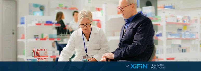 A pharmacist in a white coat consults with a customer in a suit at a pharmacy counter, with shelves of medications in the background and the XIFIN Pharmacy Solutions logo at the bottom right.